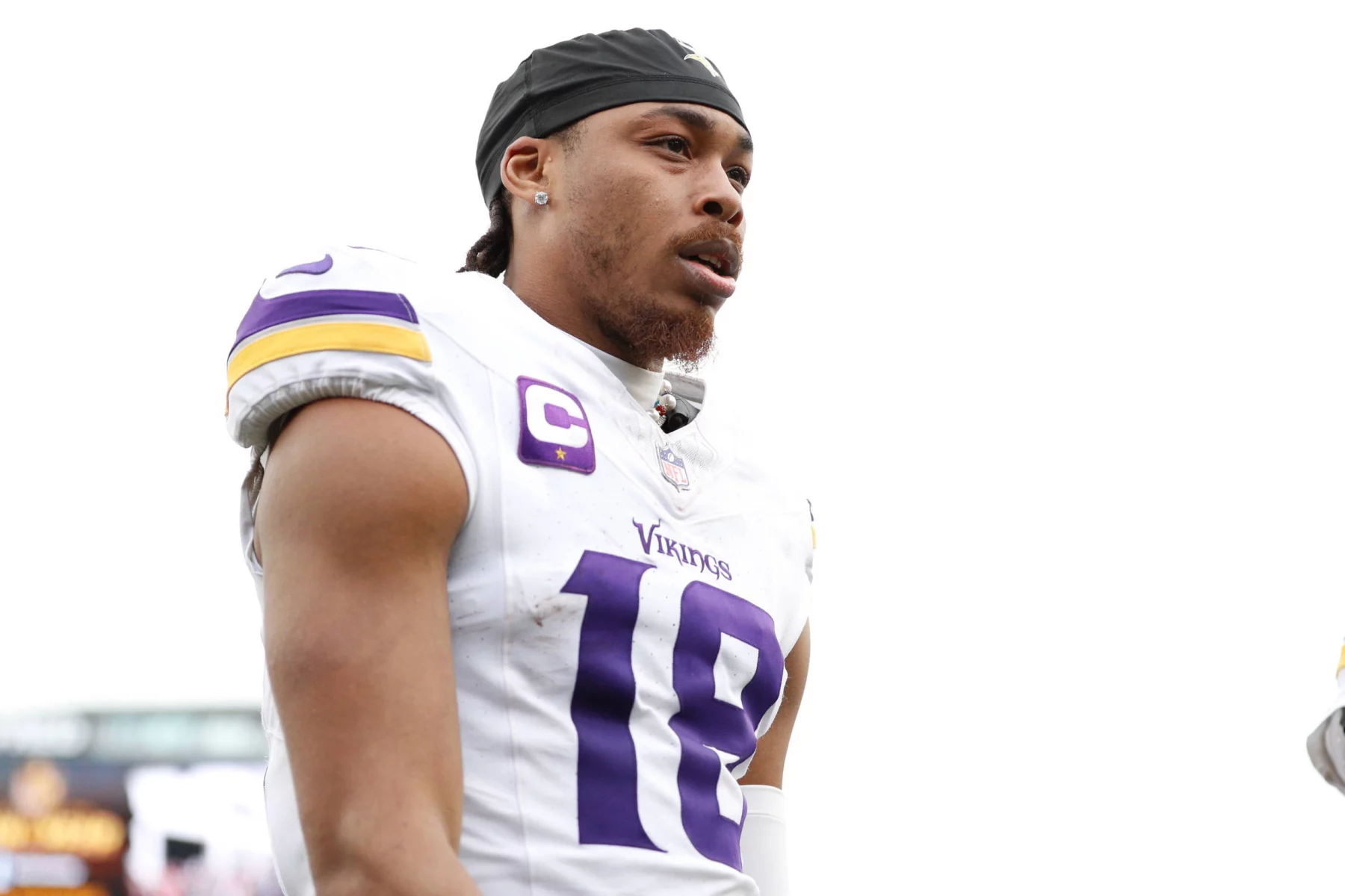 FootballR - NFL - Ein Footballspieler in einem weißen Minnesota Vikings-Trikot mit der Nummer 18 auf der Vorderseite steht auf dem Feld. Justin Jefferson, mit schwarzem Stirnband und konzentriertem Gesichtsausdruck, zeigt das Logo des Teams auf seiner Uniform. Der Hintergrund ist hell und verschwommen und fängt die Intensität seines Trainings ein. Diese Beschreibung wurde automatisch generiert.