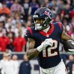 FootballR - NFL - Ein Footballspieler in der marineblau-roten Houston Texans-Uniform läuft mit einem Football in der Hand über das Feld. Er wirkt konzentriert und blickt nach vorn. Nico Collins, der sich gemäß seinem Vertrag verpflichtet fühlt, stürmt voran, während der Hintergrund mit einer Menge Zuschauer auf den Tribünen verschwimmt. Diese Beschreibung wurde automatisch generiert.