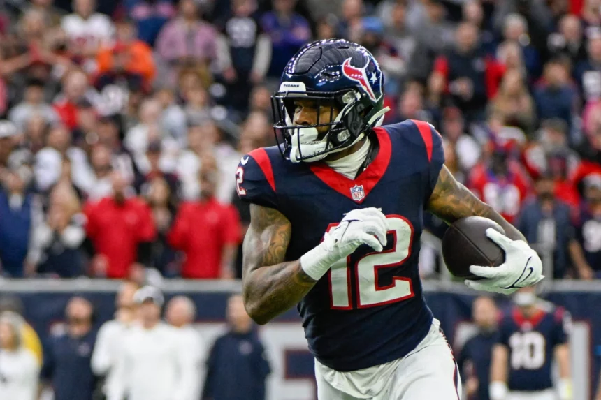 FootballR - NFL - Ein Footballspieler in der marineblau-roten Houston Texans-Uniform läuft mit einem Football in der Hand über das Feld. Er wirkt konzentriert und blickt nach vorn. Nico Collins, der sich gemäß seinem Vertrag verpflichtet fühlt, stürmt voran, während der Hintergrund mit einer Menge Zuschauer auf den Tribünen verschwimmt. Diese Beschreibung wurde automatisch generiert.