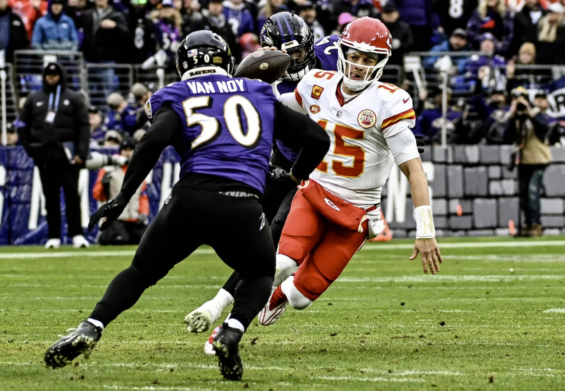 FootballR - NFL Saison 2024 - Der Quarterback der Kansas City Chiefs in einer rot-weißen Uniform weicht während eines spannenden NFL-Spiels einem Tackle eines Spielers der Baltimore Ravens im lila Trikot mit der Nummer 50 und dem Namen „Van Noy“ aus. Diese Beschreibung wurde automatisch generiert.