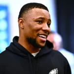 FootballR - NFL - Saquon Barkley : Ein Mann mit kurzen Haaren und Bart lächelt und schaut dabei leicht zur Seite. Er trägt einen schwarzen Kapuzenpullover mit dem Logo der Philadelphia Eagles. Der Hintergrund ist verschwommen, aber man sieht andere Leute, die stehen und reden. Die Szene scheint drinnen zu sein, möglicherweise eine Versammlung oder Veranstaltung. Diese Beschreibung wurde automatisch generiert.