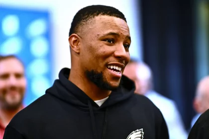 FootballR - NFL - Saquon Barkley : Ein Mann mit kurzen Haaren und Bart lächelt und schaut dabei leicht zur Seite. Er trägt einen schwarzen Kapuzenpullover mit dem Logo der Philadelphia Eagles. Der Hintergrund ist verschwommen, aber man sieht andere Leute, die stehen und reden. Die Szene scheint drinnen zu sein, möglicherweise eine Versammlung oder Veranstaltung. Diese Beschreibung wurde automatisch generiert.
