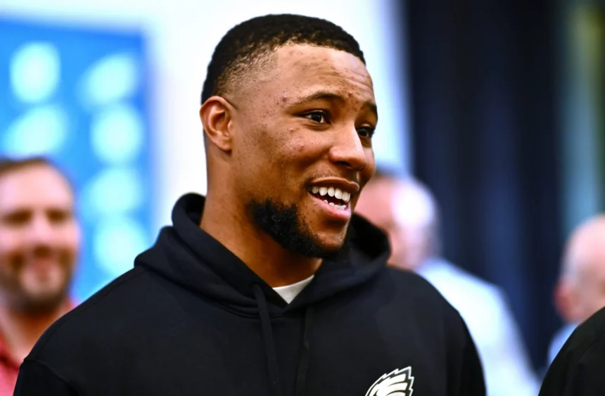 FootballR - NFL - Saquon Barkley : Ein Mann mit kurzen Haaren und Bart lächelt und schaut dabei leicht zur Seite. Er trägt einen schwarzen Kapuzenpullover mit dem Logo der Philadelphia Eagles. Der Hintergrund ist verschwommen, aber man sieht andere Leute, die stehen und reden. Die Szene scheint drinnen zu sein, möglicherweise eine Versammlung oder Veranstaltung. Diese Beschreibung wurde automatisch generiert.