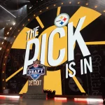Mock Draft - NFL Draft 2026 in Pittsburgh - Ein Bühnenaufbau für den NFL Draft besteht aus einem großen Bildschirm mit dem Text „The Pick is In“ und dem Logo der Pittsburgh Steelers oben. Darunter wird das NFL Draft-Logo mit „2024“ und „Detroit“ angezeigt. Helle Lichter und Kameras sind rund um die Bühne positioniert und sorgen für eine elektrisierende Atmosphäre. Diese Beschreibung wurde automatisch generiert.