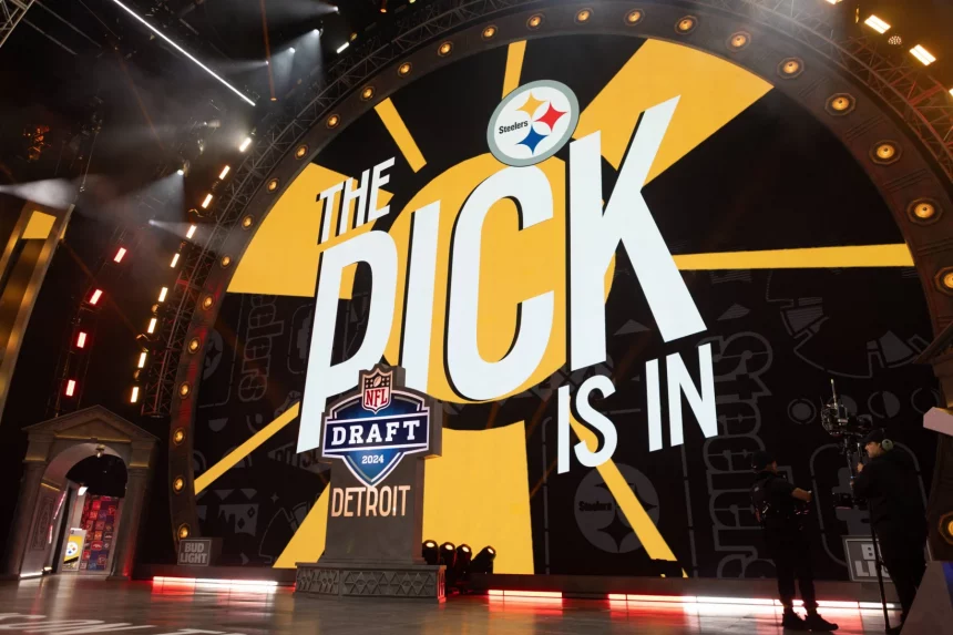 Mock Draft - NFL Draft 2026 in Pittsburgh - Ein Bühnenaufbau für den NFL Draft besteht aus einem großen Bildschirm mit dem Text „The Pick is In“ und dem Logo der Pittsburgh Steelers oben. Darunter wird das NFL Draft-Logo mit „2024“ und „Detroit“ angezeigt. Helle Lichter und Kameras sind rund um die Bühne positioniert und sorgen für eine elektrisierende Atmosphäre. Diese Beschreibung wurde automatisch generiert.
