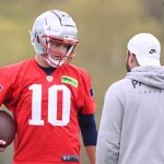 FootballR - NFL - Ein Footballspieler in einem roten Trikot mit der Nummer 10 und einem weißen Helm hält einen Football in der Hand und unterhält sich mit einem anderen Spieler, der teilweise zu sehen ist und langes, lockiges Haar hat. Sie stehen auf einem Rasenplatz. Einer von ihnen ist Drake Maye. Diese Beschreibung wurde automatisch generiert.