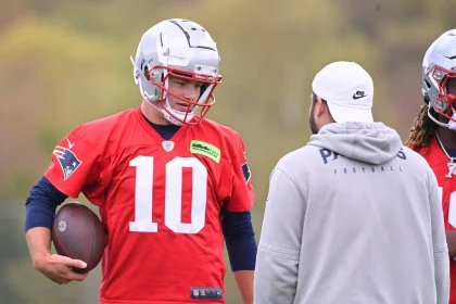 FootballR - NFL - Ein Footballspieler in einem roten Trikot mit der Nummer 10 und einem weißen Helm hält einen Football in der Hand und unterhält sich mit einem anderen Spieler, der teilweise zu sehen ist und langes, lockiges Haar hat. Sie stehen auf einem Rasenplatz. Einer von ihnen ist Drake Maye. Diese Beschreibung wurde automatisch generiert.