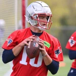 FootballR - NFL - Ein Footballspieler, Drake Maye, in einem roten Trainingstrikot und Helm, der an die Trikots von Legenden wie Jerod Mayo erinnert, bereitet sich darauf vor, einen Football zu werfen. Das Trikot trägt die Nummer 10 und das Logo der New England Patriots. Der Spieler steht auf einem Rasenfeld mit einem Tornetz im Hintergrund. Dahinter ist teilweise ein weiterer Spieler in einem roten Trikot und Helm zu sehen. Diese Beschreibung wurde automatisch generiert.