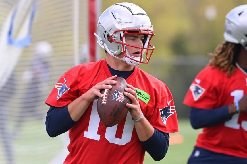 FootballR - NFL - Ein Footballspieler, Drake Maye, in einem roten Trainingstrikot und Helm, der an die Trikots von Legenden wie Jerod Mayo erinnert, bereitet sich darauf vor, einen Football zu werfen. Das Trikot trägt die Nummer 10 und das Logo der New England Patriots. Der Spieler steht auf einem Rasenfeld mit einem Tornetz im Hintergrund. Dahinter ist teilweise ein weiterer Spieler in einem roten Trikot und Helm zu sehen. Diese Beschreibung wurde automatisch generiert.