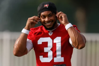 FootballR - NFL - Antoine Winfield Jr. - Ein lächelnder Footballspieler im roten Buccaneers-Trikot mit der Nummer 31 rückt seine schwarze Mütze auf einem sonnigen Übungsplatz zurecht, im Hintergrund ist verschwommenes Grün zu sehen. Diese Beschreibung wurde automatisch generiert.