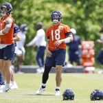 FootballR - NFL Hard Knocks - Caleb Williams - Drei Footballspieler der Chicago Bears trainieren auf einem Rasenplatz. Sie tragen orangefarbene Trikots und marineblaue Shorts. Zwei Spieler auf der linken Seite stehen und beobachten, während der Spieler auf der rechten Seite in Bewegung ist. Im Vordergrund liegen Helme auf dem Boden, während sie sich auf ihren Hard-Knocks-Moment vorbereiten. Im Hintergrund sind Bäume zu sehen. Diese Beschreibung wurde automatisch generiert.