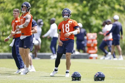 FootballR - NFL Hard Knocks - Caleb Williams - Drei Footballspieler der Chicago Bears trainieren auf einem Rasenplatz. Sie tragen orangefarbene Trikots und marineblaue Shorts. Zwei Spieler auf der linken Seite stehen und beobachten, während der Spieler auf der rechten Seite in Bewegung ist. Im Vordergrund liegen Helme auf dem Boden, während sie sich auf ihren Hard-Knocks-Moment vorbereiten. Im Hintergrund sind Bäume zu sehen. Diese Beschreibung wurde automatisch generiert.