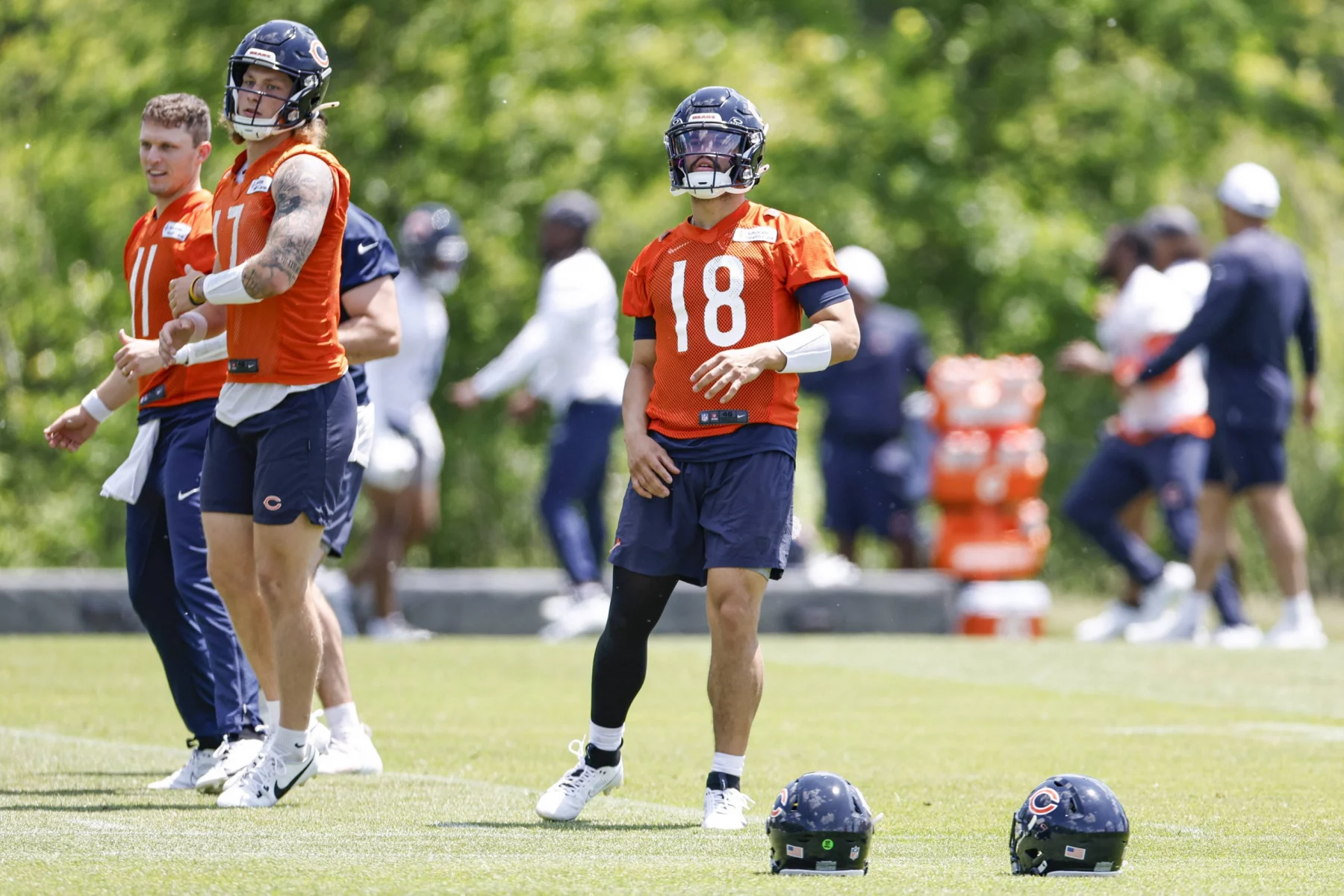 FootballR - NFL Hard Knocks - Caleb Williams - Drei Footballspieler der Chicago Bears trainieren auf einem Rasenplatz. Sie tragen orangefarbene Trikots und marineblaue Shorts. Zwei Spieler auf der linken Seite stehen und beobachten, während der Spieler auf der rechten Seite in Bewegung ist. Im Vordergrund liegen Helme auf dem Boden, während sie sich auf ihren Hard-Knocks-Moment vorbereiten. Im Hintergrund sind Bäume zu sehen. Diese Beschreibung wurde automatisch generiert.