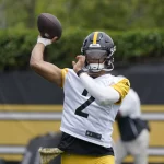 FootballR - NFL - Ein Footballspieler mit schwarzem Helm und weiß-gelbem Trikot mit der Nummer 2, der Quarterback Justin Fields ähnelt, ist mitten in einer Wurfbewegung. Er ist von einem grünen Übungsplatz im Freien umgeben, im Hintergrund ist eine weitere Person zu sehen. Die Szene scheint eine Trainings- oder Übungseinheit der Steelers zu sein. Diese Beschreibung wurde automatisch generiert.