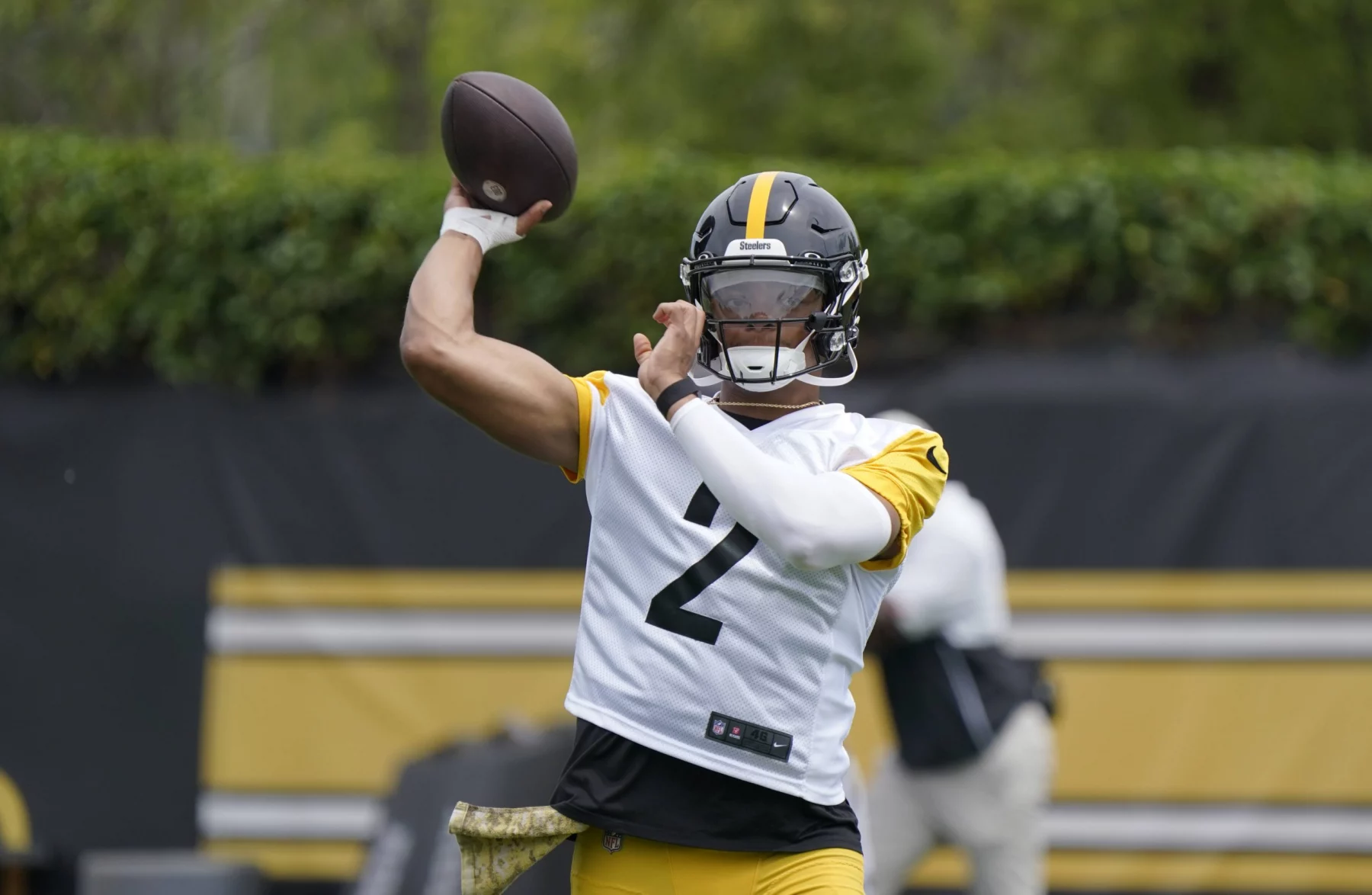 FootballR - NFL - Ein Footballspieler mit schwarzem Helm und weiß-gelbem Trikot mit der Nummer 2, der Quarterback Justin Fields ähnelt, ist mitten in einer Wurfbewegung. Er ist von einem grünen Übungsplatz im Freien umgeben, im Hintergrund ist eine weitere Person zu sehen. Die Szene scheint eine Trainings- oder Übungseinheit der Steelers zu sein. Diese Beschreibung wurde automatisch generiert.