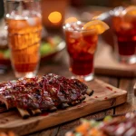 FootballR - NFL 40YARDS - Auf einem Holztisch wird ein Festmahl serviert, in der Mitte ein Schneidebrett mit glasierten BBQ-Rippchen. Um die Rippchen herum stehen Gläser mit Eistee, garniert mit Orangenscheiben. Ein unscharfer Hintergrund deutet auf andere Gerichte hin und die stimmungsvolle Beleuchtung mit Kerzen neben einem NFL-Bettwäscheset schafft eine gemütliche Essatmosphäre. Diese Beschreibung wurde automatisch generiert.