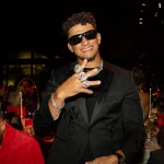 FootballR - NFL - Ein Mann in schwarzem Anzug, Sonnenbrille und mehreren großen Ringen an den Fingern lächelt und gestikuliert mit der Hand. Er steht in einem schwach beleuchteten Raum, im Hintergrund sitzen Menschen an Tischen. Ein weiterer Mann in einer roten Jacke, der sich vielleicht die Höhepunkte des Super Bowl LVIII ansieht, blickt nach links hinunter. Diese Beschreibung wurde automatisch generiert.