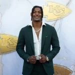 FootballR - NFL - Ein Mann in einem dunkelgrünen Anzug und einem weißen Hemd steht lächelnd vor einem Hintergrund mit mehreren Logos der Kansas City Chiefs. Er hat geflochtene Haare und trägt eine Halskette, eine Uhr und einen glänzenden Chiefs-Ring aus dem Super Bowl LVIII. Der marmorähnliche Hintergrund weist markante goldene Chiefs-Logos auf, perfekt für Bilder und Videos. Diese Beschreibung wurde automatisch generiert.