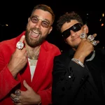 FootballR - NFL - Travis Kelce und Patrick Mahomes - Zwei Männer in stilvoller Kleidung, jeder trägt mehrere große Ringe an seinen Fingern. Der Mann links trägt eine rote Jacke, eine Goldkette und eine runde Brille, während der Mann rechts einen schwarzen Anzug, eine Silberkette und eine dunkle Sonnenbrille trägt. Beide lächeln in die Kamera, nachdem sie den Super Bowl LVIII mit den Chiefs gesehen haben. Diese Beschreibung wurde automatisch generiert.
