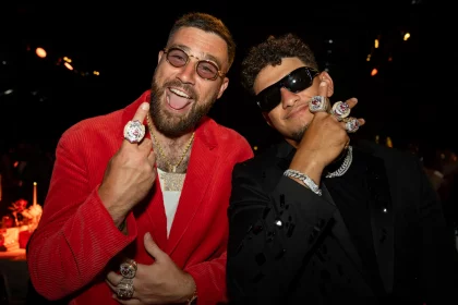 FootballR - NFL - Travis Kelce und Patrick Mahomes - Zwei Männer in stilvoller Kleidung, jeder trägt mehrere große Ringe an seinen Fingern. Der Mann links trägt eine rote Jacke, eine Goldkette und eine runde Brille, während der Mann rechts einen schwarzen Anzug, eine Silberkette und eine dunkle Sonnenbrille trägt. Beide lächeln in die Kamera, nachdem sie den Super Bowl LVIII mit den Chiefs gesehen haben. Diese Beschreibung wurde automatisch generiert.