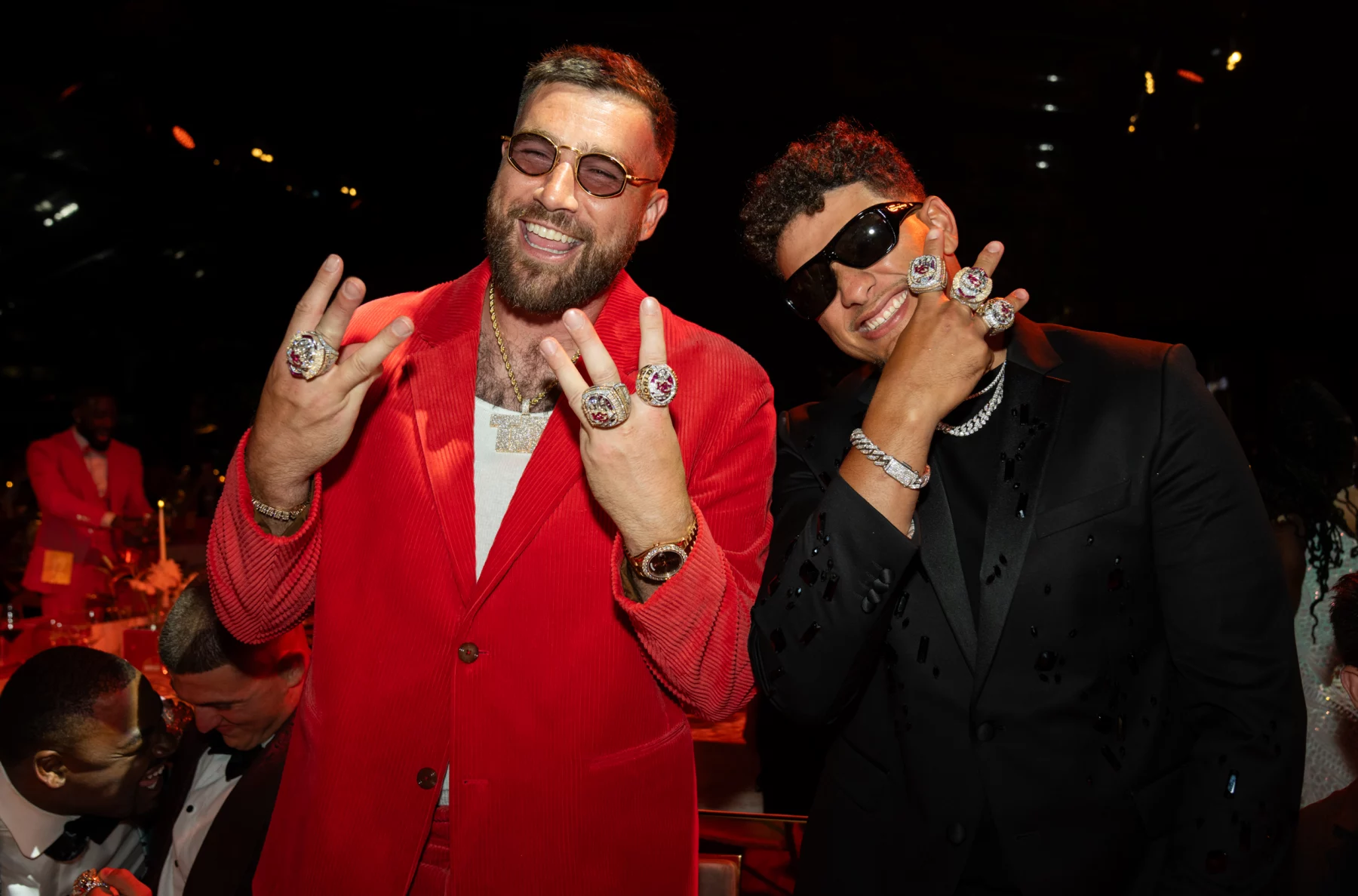 FootballR - NFL - Zwei Männer lächeln und zeigen mehrere große Ringe an ihren Händen. Der Mann links trägt einen roten Anzug und eine Sonnenbrille, während der Mann rechts einen schwarzen Anzug, eine schwarze Sonnenbrille und ein Diamantarmband trägt. Sie posieren zusammen bei einer Indoor-Veranstaltung mit anderen Teilnehmern im Hintergrund und machen Aufnahmen für Bilder & Videos. Diese Beschreibung wurde automatisch generiert.