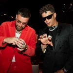 FootballR - NFL Travis Kelce und Patrick Mahomes - Zwei Männer in stilvoller Kleidung sind in einer lebhaften Umgebung zu sehen, die an die Super Bowl LVIII-Feier der Chiefs erinnert. Der Mann links trägt einen leuchtend roten Anzug mit weißem Hemd und ist mit mehreren Ringen geschmückt. Der Mann rechts trägt einen schwarzen Anzug, eine Sonnenbrille und ebenfalls mehrere Ringe, während sie ihren Bling wie wertvolle Galerie-Stücke lebhaft zur Schau stellen. Diese Beschreibung wurde automatisch generiert.
