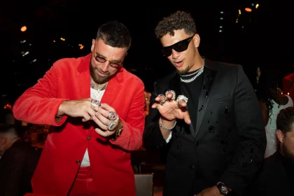 FootballR - NFL Travis Kelce und Patrick Mahomes - Zwei Männer in stilvoller Kleidung sind in einer lebhaften Umgebung zu sehen, die an die Super Bowl LVIII-Feier der Chiefs erinnert. Der Mann links trägt einen leuchtend roten Anzug mit weißem Hemd und ist mit mehreren Ringen geschmückt. Der Mann rechts trägt einen schwarzen Anzug, eine Sonnenbrille und ebenfalls mehrere Ringe, während sie ihren Bling wie wertvolle Galerie-Stücke lebhaft zur Schau stellen. Diese Beschreibung wurde automatisch generiert.