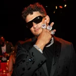 FootballR - NFL - Eine Person mit Sonnenbrille und schwarzem Anzug posiert selbstbewusst und zeigt vier große, mit Diamanten besetzte Ringe an ihren Fingern. Sie trägt eine dicke Halskette und steht in einer schwach beleuchteten Umgebung. Im verschwommenen Hintergrund sind andere Personen und Tische zu sehen, was an eine Feier nach dem Super Bowl erinnert. Diese Beschreibung wurde automatisch generiert.