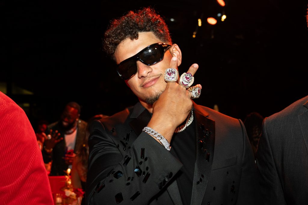 FootballR - NFL - Ein Mann mit kurzen lockigen Haaren und Sonnenbrille trägt ein schwarzes Jackett mit glänzenden schwarzen Mustern. Er zeigt drei große, mit Juwelen besetzte Ringe an seiner rechten Hand, indem er sie vor sein Gesicht hält und die Finger so spreizt, dass die Ringe zur Geltung kommen. Der schwach beleuchtete Hintergrund erinnert an eine lebhafte Galerie während der Feierlichkeiten zum Super Bowl LVIII. Diese Beschreibung wurde automatisch generiert.