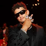 FootballR - NFL - Ein Mann mit kurzen lockigen Haaren und Sonnenbrille trägt ein schwarzes Jackett mit glänzenden schwarzen Mustern. Er zeigt drei große, mit Juwelen besetzte Ringe an seiner rechten Hand, indem er sie vor sein Gesicht hält und die Finger so spreizt, dass die Ringe zur Geltung kommen. Der schwach beleuchtete Hintergrund erinnert an eine lebhafte Galerie während der Feierlichkeiten zum Super Bowl LVIII. Diese Beschreibung wurde automatisch generiert.