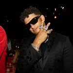 FootballR - NFL - Eine Person in einem schwarzen Anzug und mit schwarzer Sonnenbrille posiert vor einem dunklen Hintergrund, was an die Feierlichkeiten zum Super Bowl LVIII erinnert. Sie hält ihre Hand hoch und zeigt mehrere große, glitzernde Ringe. Die Person hat kurzes lockiges Haar und ist von anderen umgeben, darunter jemand in einer roten Chiefs-Jacke zu ihrer Linken und eine weitere Person, die zu ihrer Rechten teilweise sichtbar ist. Diese Beschreibung wurde automatisch generiert.