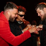 FootballR - NFL - Drei Männer lächeln und schauen auf ihre Hände, die mit großen, auffälligen Chiefs-Ringen aus dem Super Bowl LVIII geschmückt sind. Ein Mann trägt eine rote Jacke und eine Brille, der mittlere Mann trägt ein schwarzes Outfit mit Sonnenbrille und der dritte Mann trägt einen dunklen Anzug mit Krawatte. Der Hintergrund ist schwach beleuchtet und suggeriert ein festliches oder feierliches Ereignis, das sich für Bilder und Videos eignet. Diese Beschreibung wurde automatisch generiert.