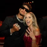 FootballR - NFL - Ein Mann und eine Frau posieren fröhlich in einem Innenbereich bei einer scheinbar formellen Veranstaltung. Der Mann trägt eine Sonnenbrille und einen mit Schmuck geschmückten schwarzen Anzug und hält stolz vier große Super Bowl LVIII-Ringe hoch. Die Frau in einem roten Kleid mit Fransen zeigt ebenfalls Ringe. Beide lächeln breit. Neben ihnen steht ein weiterer Mann in einem Anzug von Galerie. Diese Beschreibung wurde automatisch generiert.