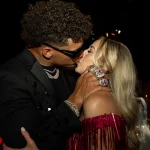 FootballR - NFL - Ein Mann in schwarzem Anzug und Sonnenbrille küsst leidenschaftlich eine Frau in einem schulterfreien, glänzenden roten Fransenkleid. Sie stehen eng beieinander in einem schwach beleuchteten Raum mit verschwommenen Figuren und leuchtenden Lichtern und erzeugen eine elektrisierende Partyatmosphäre wie beim Super Bowl LVIII. Diese Beschreibung wurde automatisch generiert.