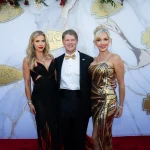 FootballR - NFL - Drei Personen stehen zusammen auf einem roten Teppich vor einem weißen Marmorhintergrund mit goldenen Logos der Kansas City Chiefs. Der Mann in der Mitte trägt einen schwarzen Smoking mit goldener Fliege, die Frau links ein schwarzes Kleid mit roten Akzenten und die Frau rechts ein gold-schwarz gestreiftes Kleid. Sie waren anwesend, um den jüngsten Super Bowl der Chiefs zu feiern. Diese Beschreibung wurde automatisch generiert.