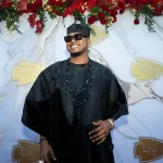 FootballR - NFL - Ein Mann mit einer traditionellen schwarzen Agbada, dunkler Sonnenbrille und passender Kappe steht lächelnd da. Er ist mit mehreren schwarzen Perlenketten geschmückt. Hinter ihm ist ein Hintergrund mit roten Rosen und Logos der Kansas City Chiefs auf einem Marmordesign zu sehen, das Galeries Tribut an den Super Bowl LVIII feiert. Diese Beschreibung wurde automatisch generiert.