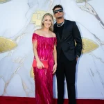 FootballR - NFL - Ein Paar posiert auf einem roten Teppich vor einem weiß-goldenen Hintergrund, perfekt für Bilder und Videos. Die Frau glänzt in einem schulterfreien, mit Fransen verzierten, metallisch-rosa Kleid und hält eine rote Clutch in der Hand. Der Mann steht hoch erhoben in einem verzierten schwarzen Anzug mit passender Kleidung und glänzenden Schuhen. Beide lächeln strahlend in die Kamera. Diese Beschreibung wurde automatisch generiert.