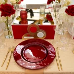 FootballR - NFL - Ein elegant gedeckter Tisch mit roten Glastellern, goldenem Besteck und durchsichtigen Weingläsern auf einer beigen Tischdecke. Rote Rosen und weiße Blumen in schmalen Vasen ergänzen ein verspiegeltes Tafelaufsatz. Dekorative Karten mit roten und goldenen Akzenten spiegeln die Pracht der Super Bowl LVIII-Ringfeier der Chiefs wider.
 Diese Beschreibung wurde automatisch generiert.