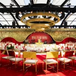 FootballR - NFL - Ein elegantes Event-Setup für die Ringzeremonie der Kansas City Chiefs umfasst eine atemberaubende Bühne mit einem rot-goldenen Hintergrund, auf dem das Logo des Teams und das Logo des Super Bowl LVIII zu sehen sind. Goldene Stühle umgeben weiße Tische, die mit Blumen und Kerzen auf einem plüschigen roten Teppich geschmückt sind, alles unter einem durchsichtigen, kuppelartigen Baldachin mit Beleuchtungskörpern. Diese Beschreibung wurde automatisch generiert.