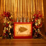 FootballR - NFL - Vier Trophäen von Vince Lombardi werden in einer Glasvitrine ausgestellt, die auf einem Marmorsockel mit einem Logo der Kansas City Chiefs steht. Die Trophäen sind von hohen, eleganten Blumenarrangements mit einer Mischung aus roten, weißen und goldenen Blumen umgeben. Ein goldener Vorhang dient als Hintergrund – eine perfekte Kulisse für Ihre Bilder- und Videosammlung. Diese Beschreibung wurde automatisch generiert.