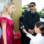 FootballR - NFL - Eine Frau in einem leuchtend rosa Kleid mit Fransen hält eine rote Clutch und lächelt, während sie von einem Kind mit einem Mikrofon interviewt wird. Ein schwarz gekleideter Mann mit Sonnenbrille und mehrreihigen Halsketten, der einen Chiefs-Ring aus dem Super Bowl LVIII trägt, beobachtet das Ganze. Sie stehen auf einem roten Teppich mit Grünpflanzen im Hintergrund und Zuschauern in der Nähe. Diese Beschreibung wurde automatisch generiert.