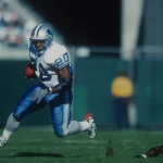 FootballR - NFL - Ein Footballspieler in weiß-blauer Uniform läuft mit dem Ball über eine Wiese und imitiert Barry Sanders, während er einem gegnerischen Spieler in schwarzer Uniform ausweicht, der fällt. Der Spieler mit dem Ball trägt die Nummer 20 und einen blauen Helm. Im Hintergrund sind verschwommene Tribünen und eine Person in einem gelben Trikot zu sehen. Diese Beschreibung wurde automatisch generiert.