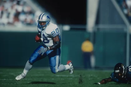 FootballR - NFL - Ein Footballspieler in weiß-blauer Uniform läuft mit dem Ball über eine Wiese und imitiert Barry Sanders, während er einem gegnerischen Spieler in schwarzer Uniform ausweicht, der fällt. Der Spieler mit dem Ball trägt die Nummer 20 und einen blauen Helm. Im Hintergrund sind verschwommene Tribünen und eine Person in einem gelben Trikot zu sehen. Diese Beschreibung wurde automatisch generiert.