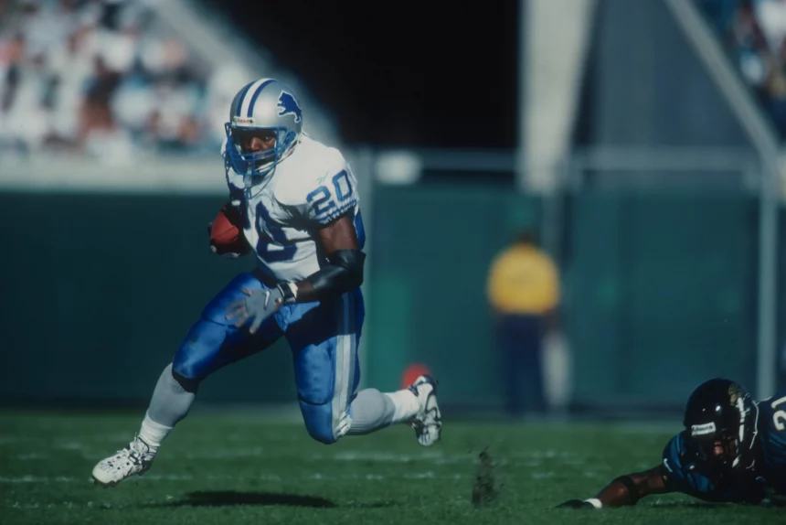 FootballR - NFL - Ein Footballspieler in weiß-blauer Uniform läuft mit dem Ball über eine Wiese und imitiert Barry Sanders, während er einem gegnerischen Spieler in schwarzer Uniform ausweicht, der fällt. Der Spieler mit dem Ball trägt die Nummer 20 und einen blauen Helm. Im Hintergrund sind verschwommene Tribünen und eine Person in einem gelben Trikot zu sehen. Diese Beschreibung wurde automatisch generiert.