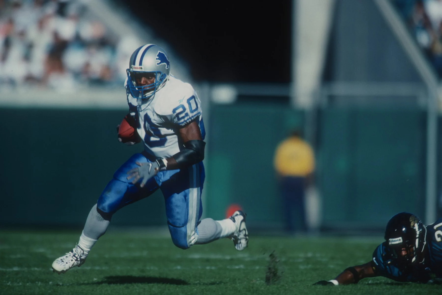 FootballR - NFL - Ein Footballspieler in weiß-blauer Uniform läuft mit dem Ball über eine Wiese und imitiert Barry Sanders, während er einem gegnerischen Spieler in schwarzer Uniform ausweicht, der fällt. Der Spieler mit dem Ball trägt die Nummer 20 und einen blauen Helm. Im Hintergrund sind verschwommene Tribünen und eine Person in einem gelben Trikot zu sehen. Diese Beschreibung wurde automatisch generiert.