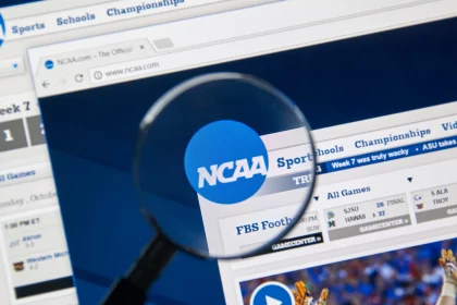 College Footballer - NFL - Eine Lupe konzentriert sich auf das NCAA Logo, das auf einem Computerbildschirm angezeigt wird, und hebt in den Browser-Tabs verschiedene Abschnitte wie „Sport“, „Schulen“ und „Meisterschaften“ hervor. Unter dem Logo gibt es einen Zeitplan für Footballspiele und ein Video-Thumbnail mit einem Spieler in orange-weißer Uniform, was die umfangreichen Kommerzialisierungsbemühungen der NCAA hervorhebt. Diese Beschreibung wurde automatisch generiert.