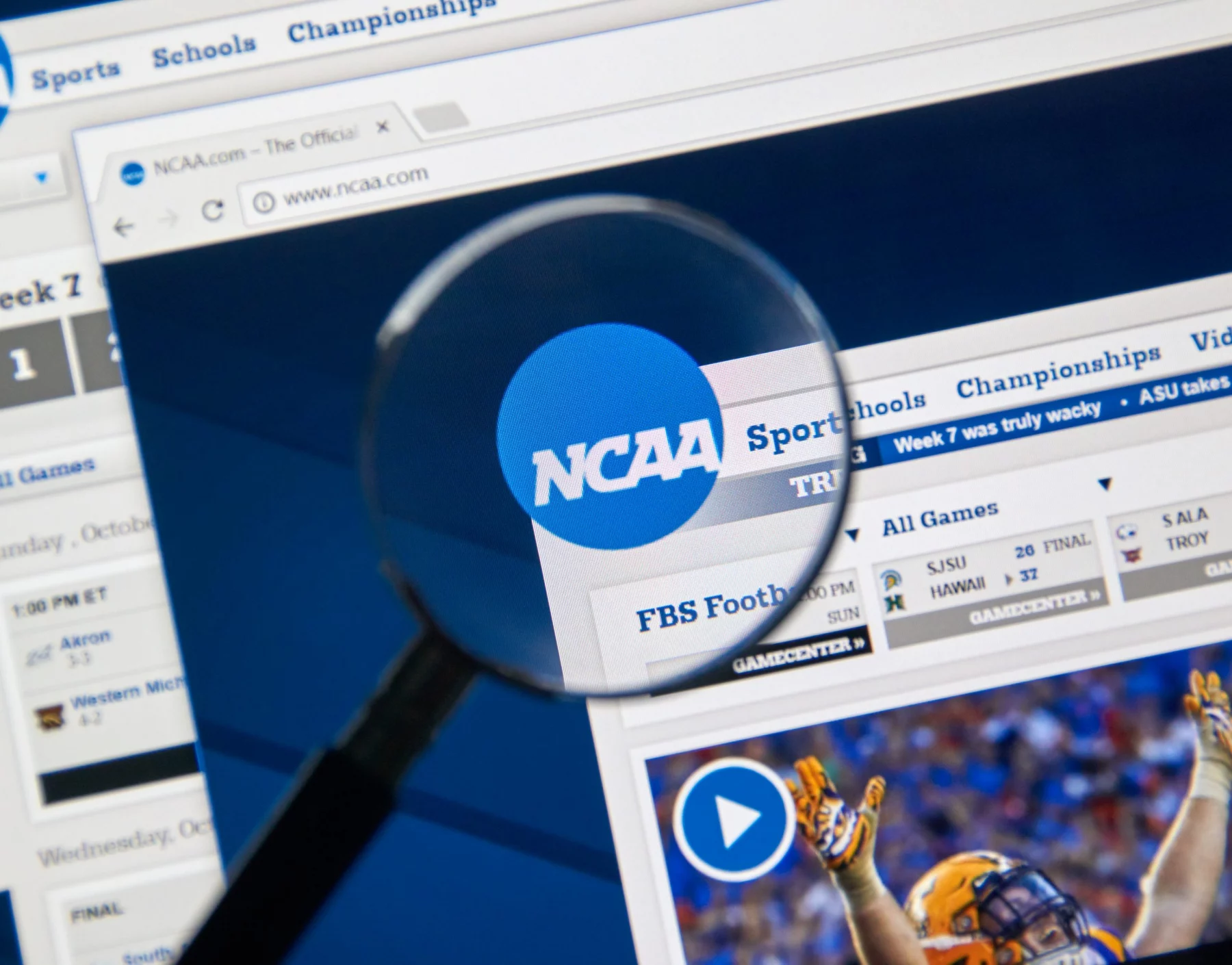 College Footballer - NFL - Eine Lupe konzentriert sich auf das NCAA Logo, das auf einem Computerbildschirm angezeigt wird, und hebt in den Browser-Tabs verschiedene Abschnitte wie „Sport“, „Schulen“ und „Meisterschaften“ hervor. Unter dem Logo gibt es einen Zeitplan für Footballspiele und ein Video-Thumbnail mit einem Spieler in orange-weißer Uniform, was die umfangreichen Kommerzialisierungsbemühungen der NCAA hervorhebt. Diese Beschreibung wurde automatisch generiert.