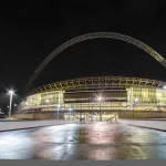 FootballR - NFL Jacksonville Jaguars - Das Bild zeigt das Wembley-Stadion in London, das nachts beleuchtet ist und über dem sich ein markanter Bogen befindet. Die Glasfassade des Stadions leuchtet warm, während die Umgebung nur schwach beleuchtet ist. Ein nasser, reflektierender Weg führt zum Haupteingang des Stadions und trägt zu der ruhigen nächtlichen Atmosphäre bei, die oft während der Heimspiele der Jacksonville Jaguars herrscht. Diese Beschreibung wurde automatisch generiert.