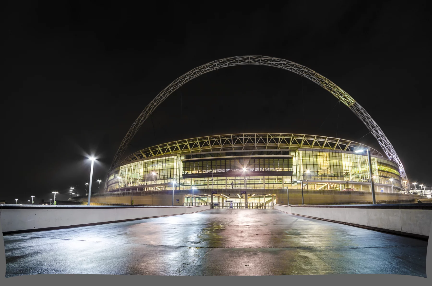 FootballR - NFL Jacksonville Jaguars - Das Bild zeigt das Wembley-Stadion in London, das nachts beleuchtet ist und über dem sich ein markanter Bogen befindet. Die Glasfassade des Stadions leuchtet warm, während die Umgebung nur schwach beleuchtet ist. Ein nasser, reflektierender Weg führt zum Haupteingang des Stadions und trägt zu der ruhigen nächtlichen Atmosphäre bei, die oft während der Heimspiele der Jacksonville Jaguars herrscht. Diese Beschreibung wurde automatisch generiert.