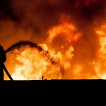 FootballR - NFL - Randall Cobb - Im Vordergrund ist die Silhouette eines Feuerwehrmanns zu sehen, der Wasser aus einem Schlauch auf einen lodernden Brand sprüht. Der Hintergrund wird von großen, leuchtend orangefarbenen Flammen und dichtem Rauch vor einem dunklen Nachthimmel dominiert, was das Ausmaß des Feuers verdeutlicht. Diese Beschreibung wurde automatisch generiert.